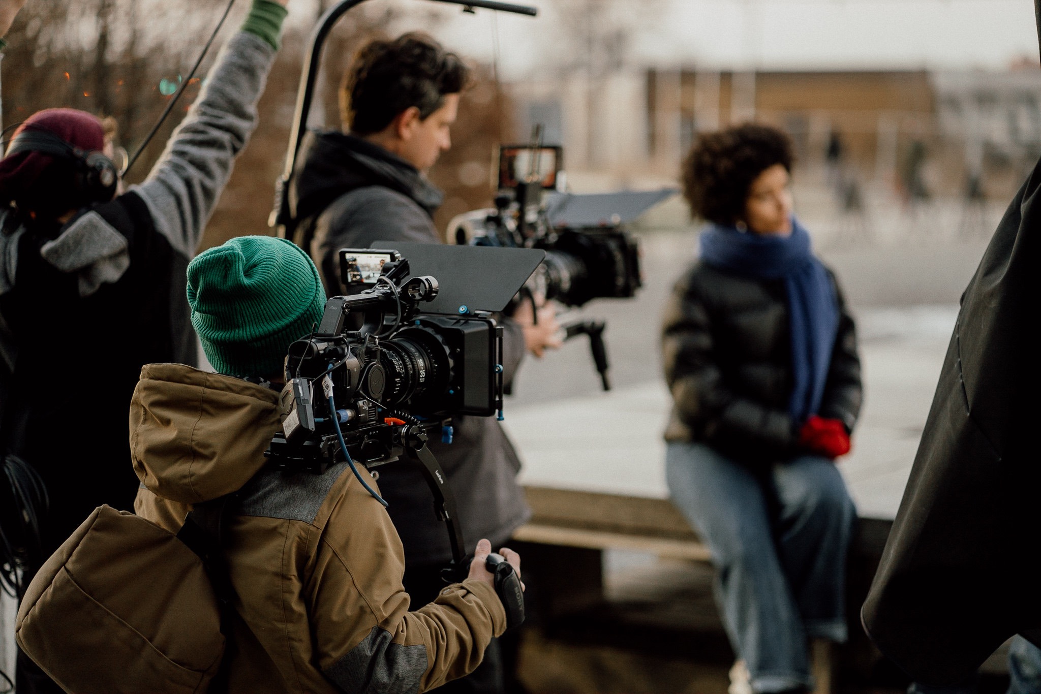 Ekipa filmowa podczas kręcenia sceny plenerowej z aktorką w niebieskim szalu i czerwonych rękawiczkach siedzącą na ławce, operator z zieloną czapką obsługuje kamerę, oświetleniowiec z mikrofonem...
