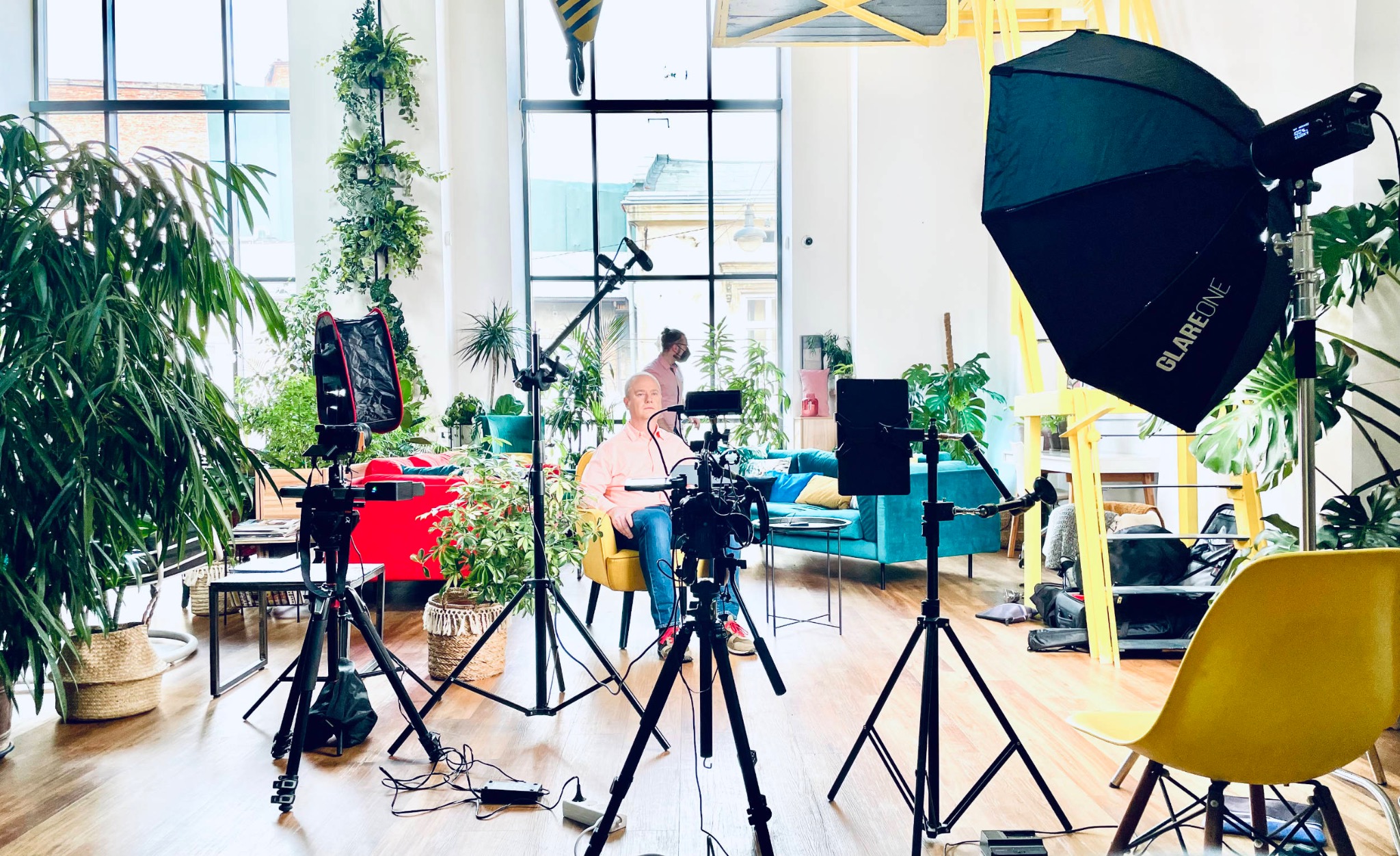 Plan filmowy w jasnym, przestronnym wnętrzu: mężczyzna siedzi na żółtym krześle przed kamerą, otoczony roślinami, lampami i rekwizytami.