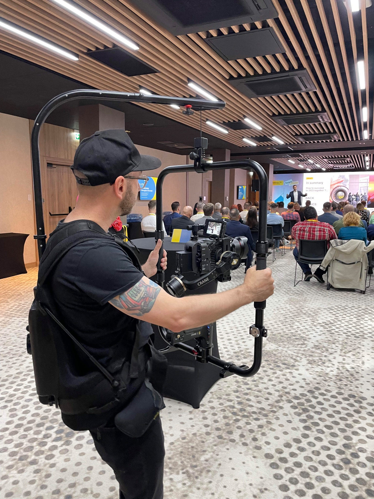 Operator kamery z tatuażem na ramieniu używa stabilizatora Crane 3S podczas filmowania prezentacji na konferencji, widoczny tłum słuchaczy i ekran z napisem 'In summary'.