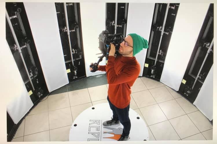 Młody mężczyzna w okularach i zielonej czapce obsługuje profesjonalną kamerę w studiu z oświetleniem panelowym, stojąc na obrotowej platformie.