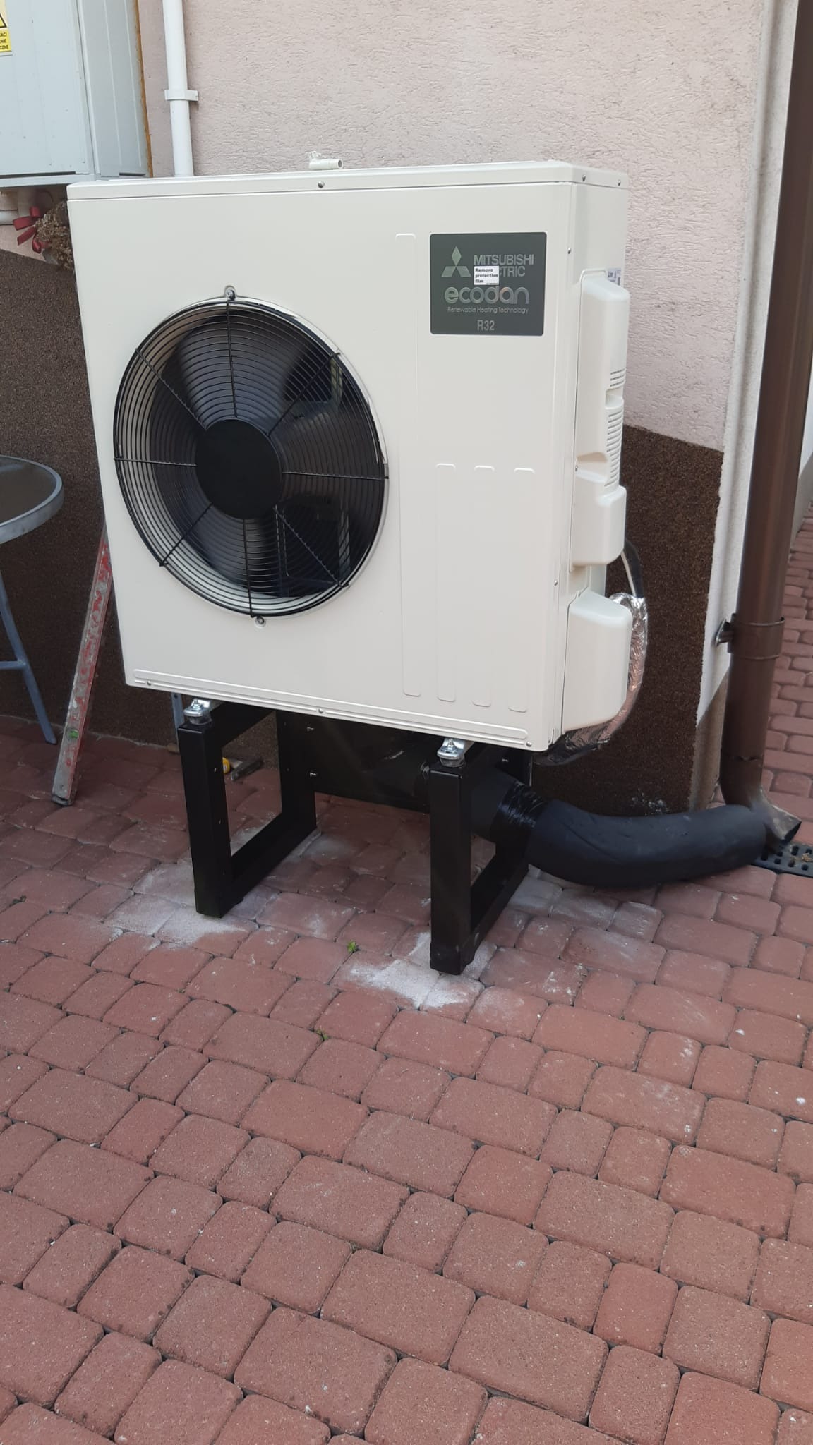 Biała pompa ciepła Mitsubishi Ecodan R32 zamontowana na czarnych metalowych wspornikach na czerwonej kostce brukowej, z widoczną izolowaną rurą odprowadzającą.
