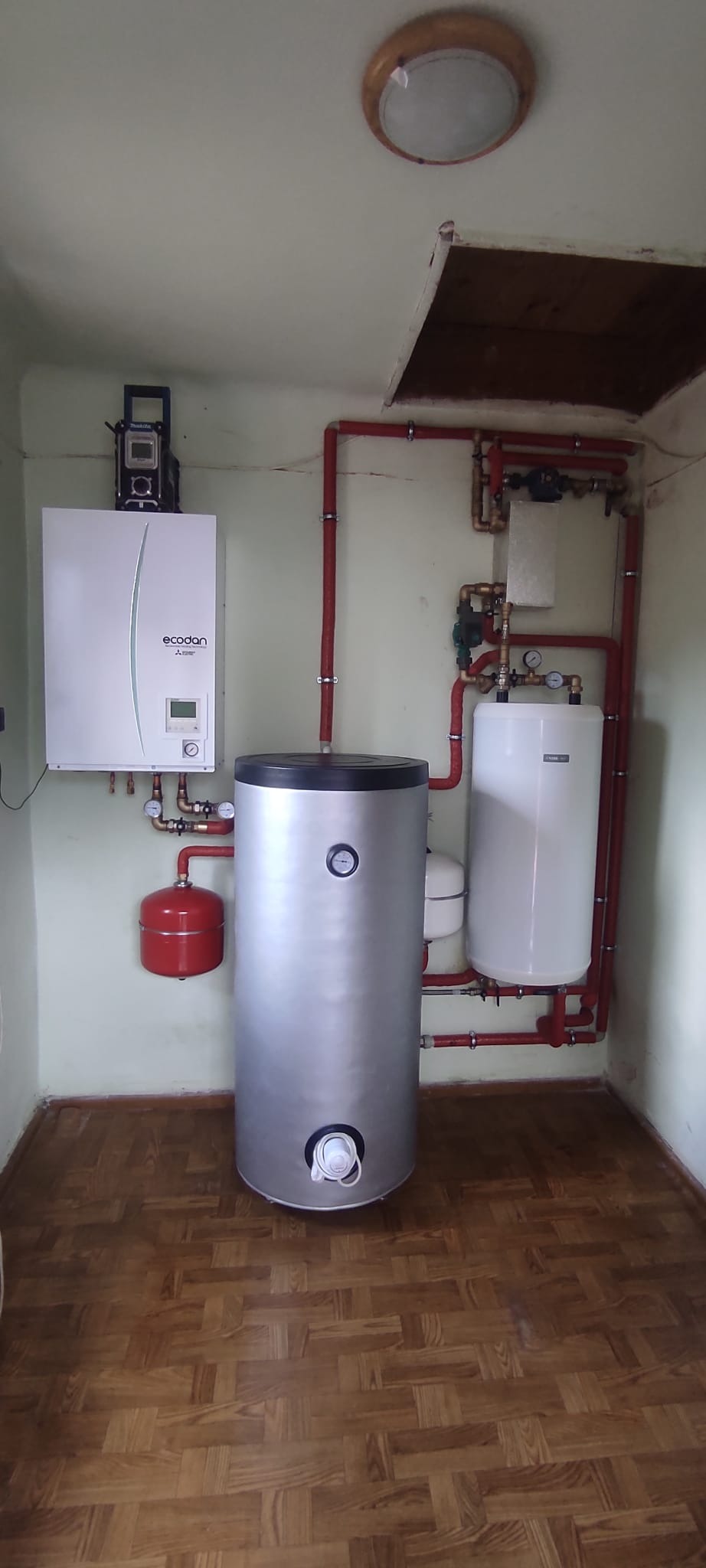 Wnętrze kotłowni z zamontowanym piecem gazowym Ecodan, srebrnym zasobnikiem ciepłej wody i czerwonym naczyniem przeponowym, instalacja z rur w kolorze czerwonym, drewniana podłoga parkietowa.
