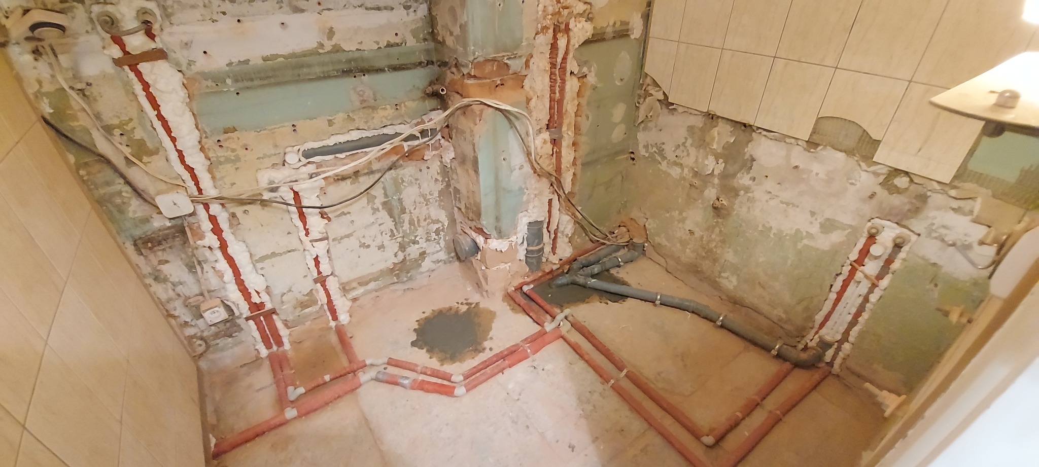 Wnętrze łazienki w trakcie remontu, widoczne nowe, czerwone rury instalacji wodnej i szare rury kanalizacyjne na tle obdrapanych ścian i częściowo zdjętych płytek.