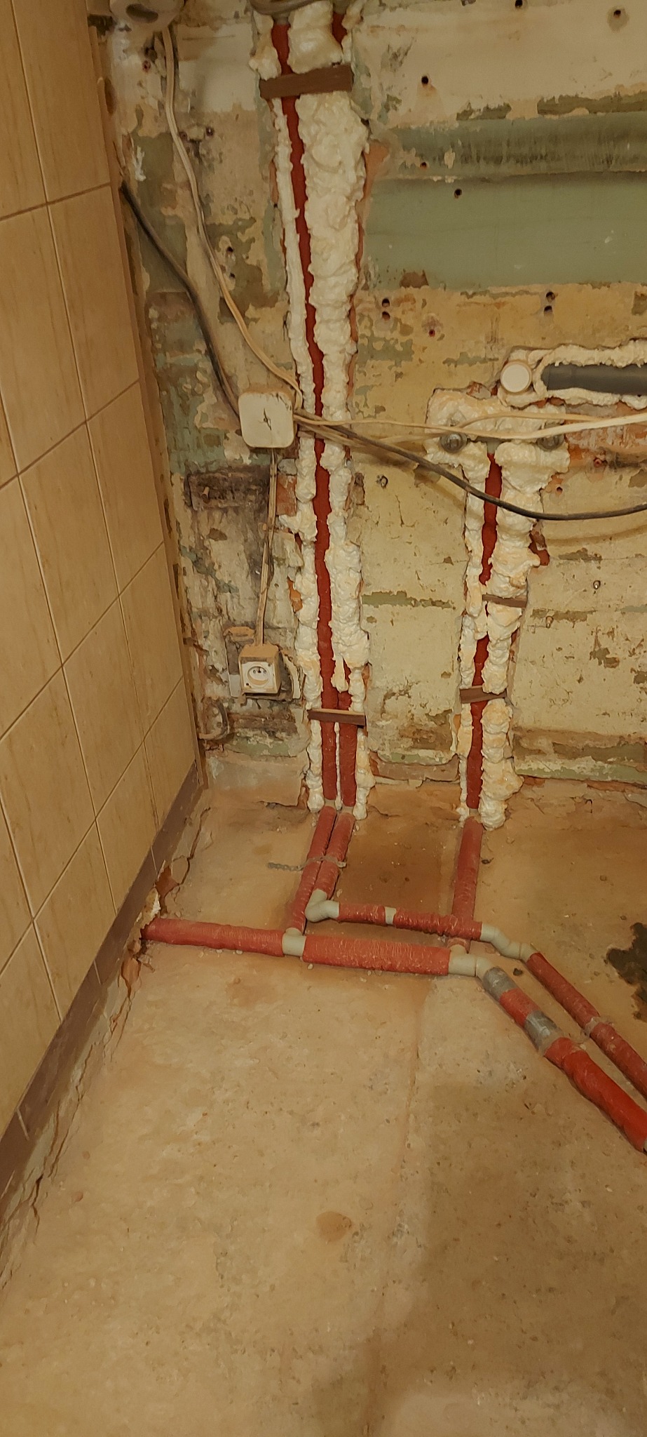 Czerwone rury instalacji centralnego ogrzewania biegnące wzdłuż ściany pokrytej pianką izolacyjną, wystające spod podłogi w pomieszczeniu w trakcie remontu, z widocznymi starymi gniazdkami...