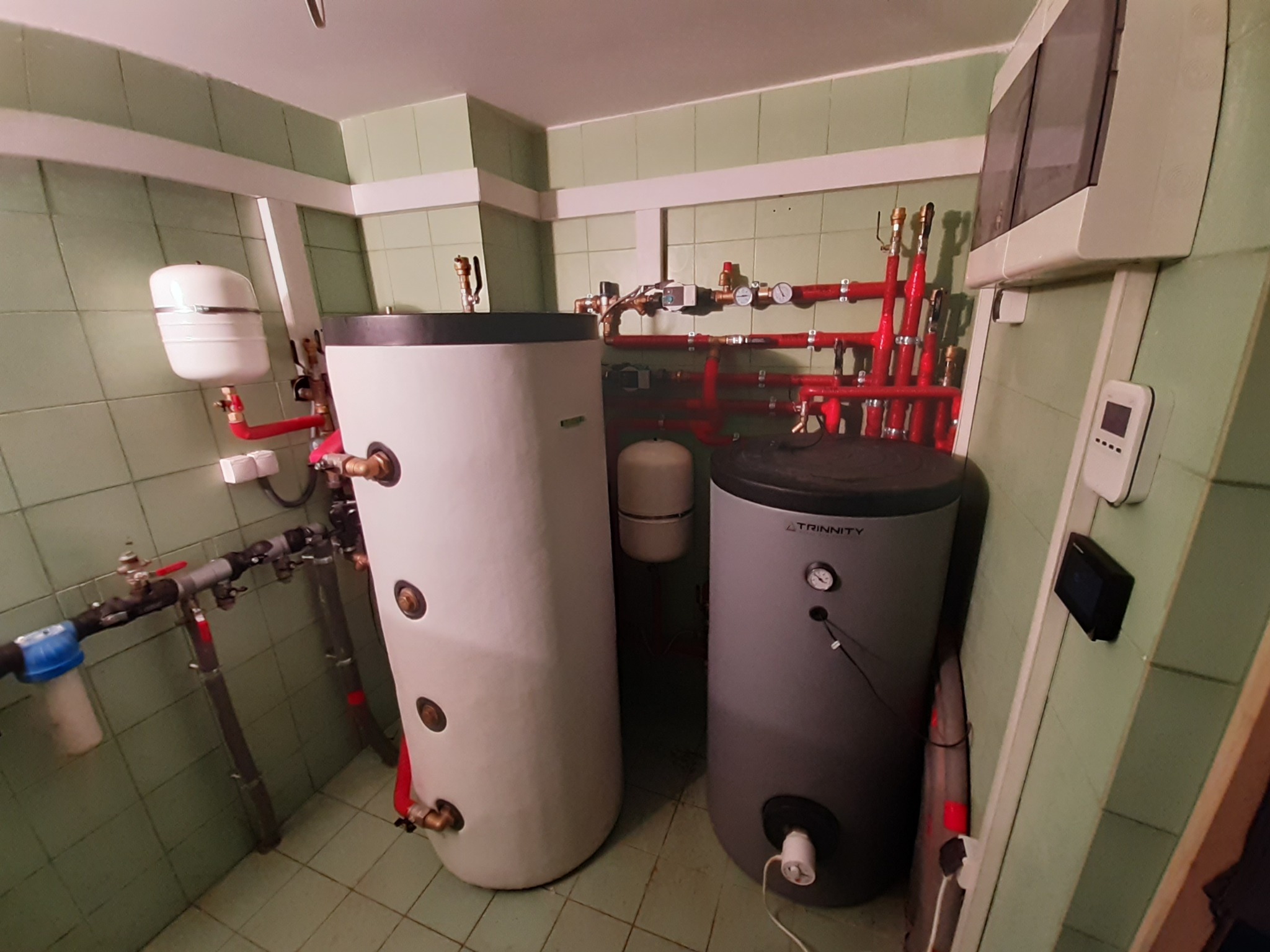 Dwa zbiorniki akumulacyjne ciepła (biały i szary) w kotłowni z zielonymi kafelkami, z widoczną instalacją hydrauliczną w kolorze czerwonym i białym, manometrami i zaworami.