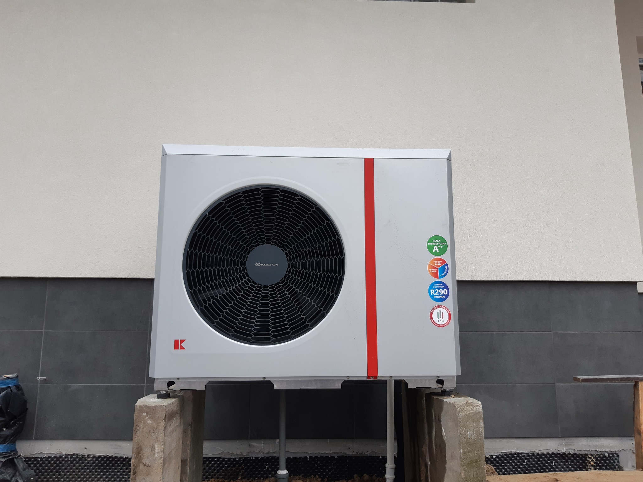 Biała pompa ciepła marki Kolton z czerwoną pionową linią, zainstalowana na betonowych podstawach przy szarej ścianie budynku z widoczną izolacją fundamentów.