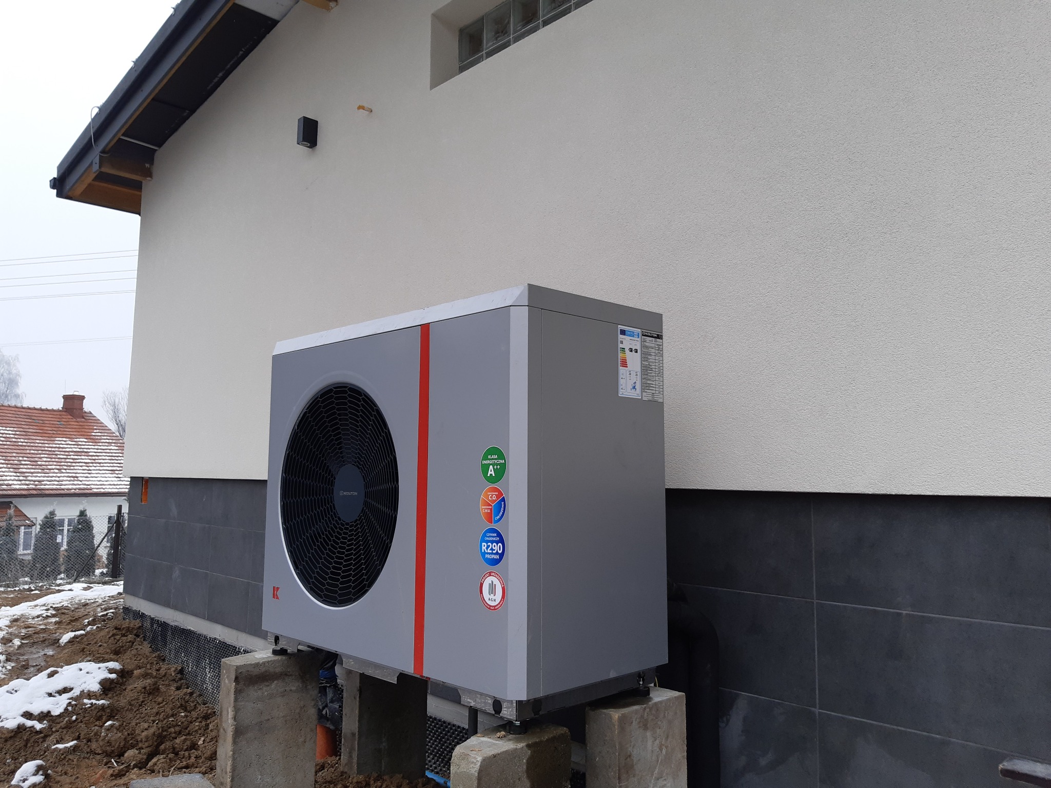 Szara pompa ciepła na betonowych podstawach, zamontowana przy elewacji domu z widocznymi naklejkami informacyjnymi o klasie energetycznej i czynniku chłodniczym R290.