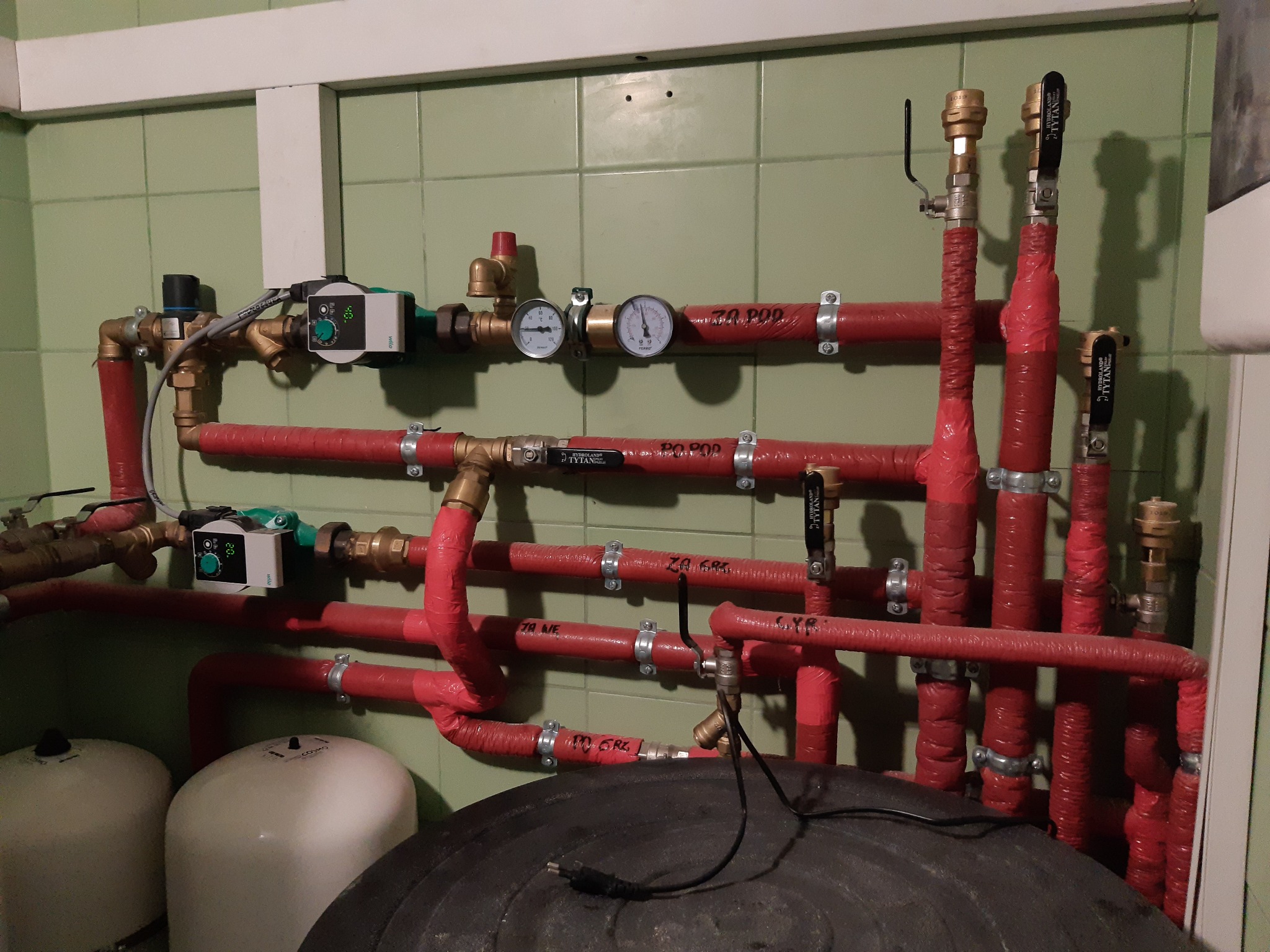 Złożona instalacja grzewcza z czerwonymi rurami, manometrami, zaworami i pompami obiegowymi w pomieszczeniu z zielonymi kafelkami.