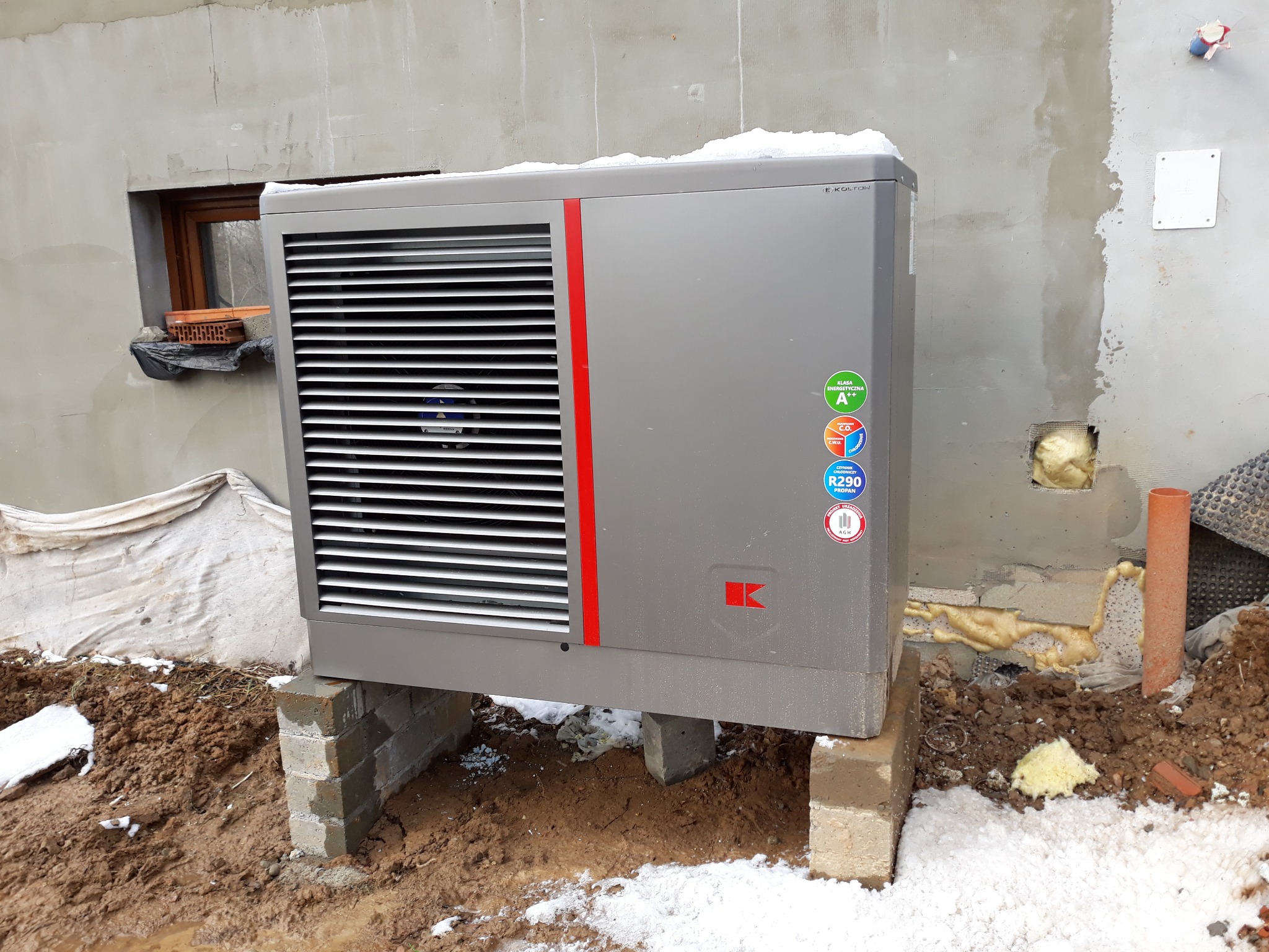 Szara pompa ciepła z czerwoną wstawką, umieszczona na betonowych blokach przed surową ścianą budynku z widocznym oknem i elementami instalacji.