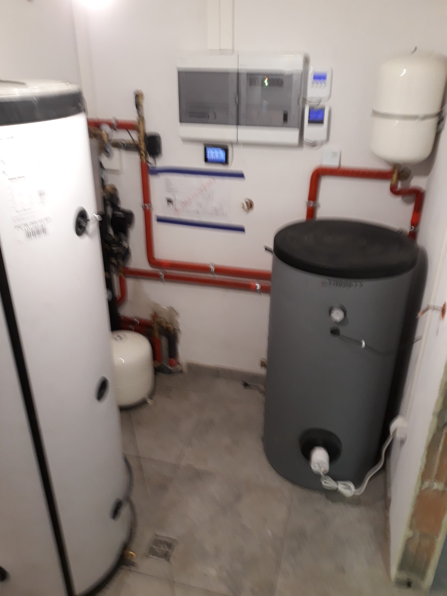 Dwa zbiorniki akumulacyjne, biały i szary, w kotłowni z widocznymi rurami instalacji grzewczej, sterownikiem i naczyniem przeponowym.