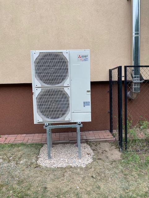 Zewnętrzna jednostka pompy ciepła Mitsubishi, zamontowana na metalowym stelażu i podsypana białym żwirem przy ścianie budynku z rurą odprowadzającą spaliny.