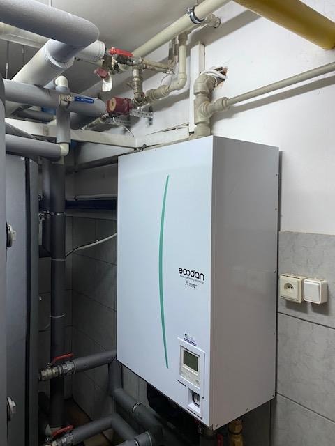 Biały piec CO marki Ecodan Mitsubishi z zielonym paskiem, zamontowany w kotłowni, widoczne rury instalacji grzewczej i gniazdka elektryczne na ścianie z szarymi płytkami.