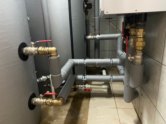 Szare rury instalacji grzewczej z zaworami, izolowane termicznie, zamontowane przy zbiornikach i ścianie z płytek ceramicznych.