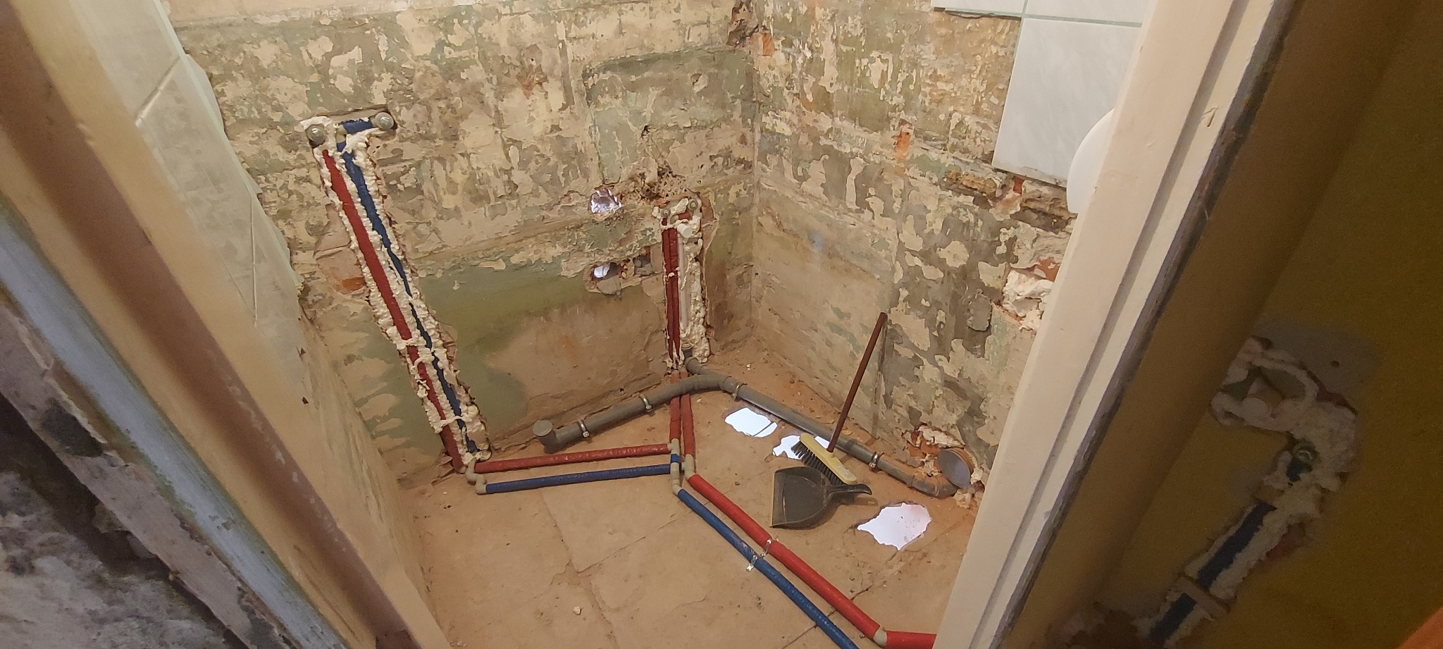 Surowy stan łazienki w trakcie remontu: widoczne rury instalacji wodnej zabezpieczone pianką montażową, szare rury kanalizacyjne, ściany z resztkami starej farby i płytek, na podłodze zmiotka...