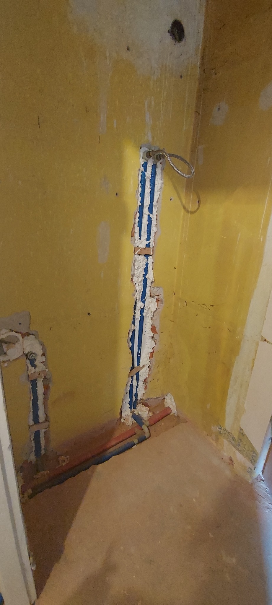 Wnęka w żółtej ścianie z nowymi, niebieskimi rurami instalacji wodnej zalane pianką montażową, widoczne fragmenty starej instalacji i surowego tynku.
