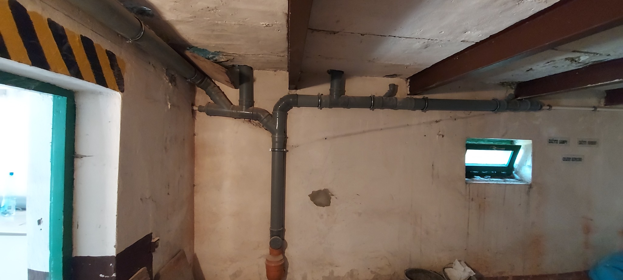 Szare rury kanalizacyjne z licznymi złączkami zamontowane pod sufitem w piwnicy z małym oknem i żółto-czarnymi pasami ostrzegawczymi na framudze drzwi.