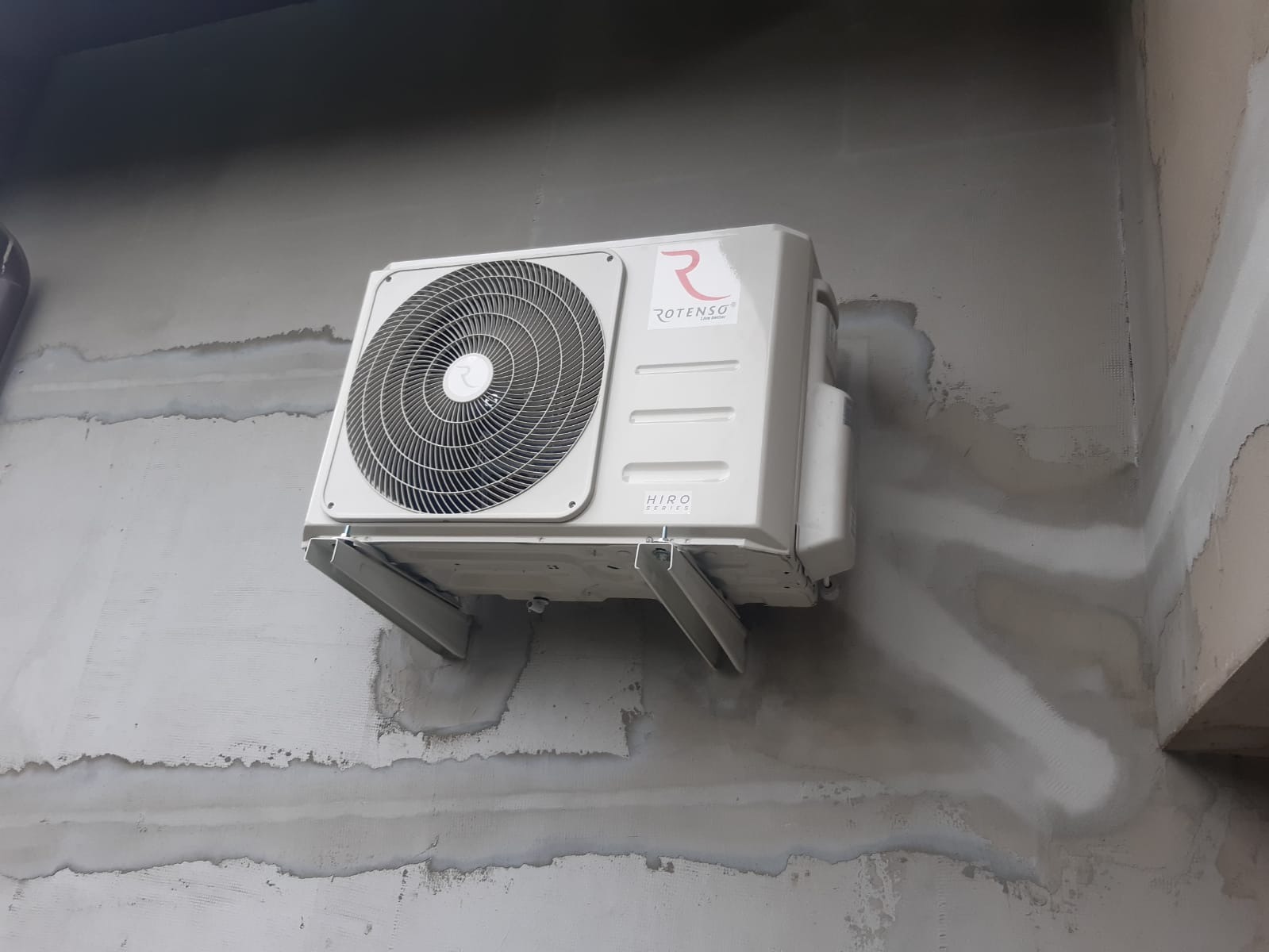 Biała jednostka zewnętrzna klimatyzacji marki Rotenso zamontowana na szarej, surowej ścianie z widocznymi śladami po pracach budowlanych.