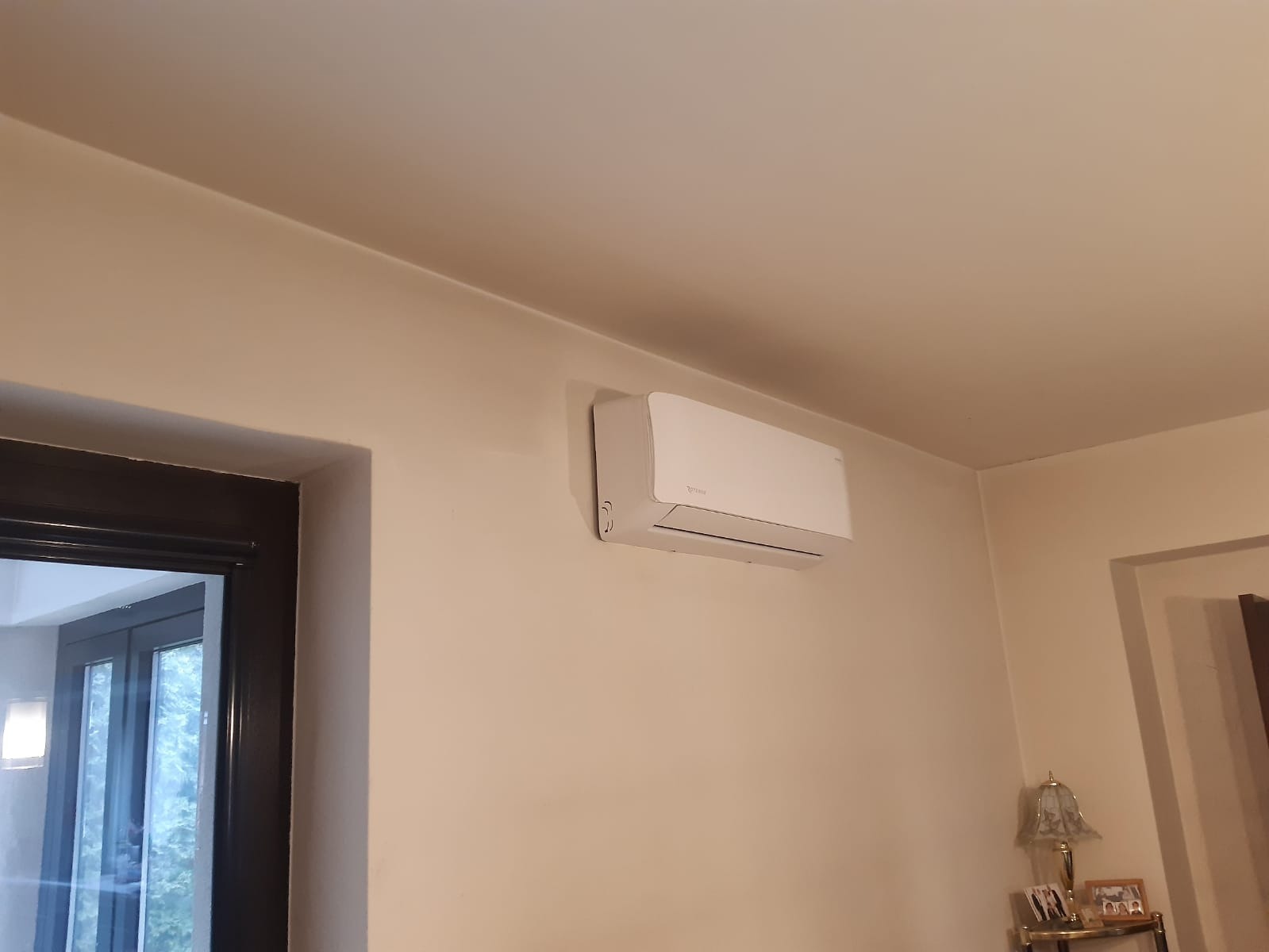 Biały klimatyzator zamontowany na beżowej ścianie w pokoju, widoczny fragment okna i lampki nocnej.