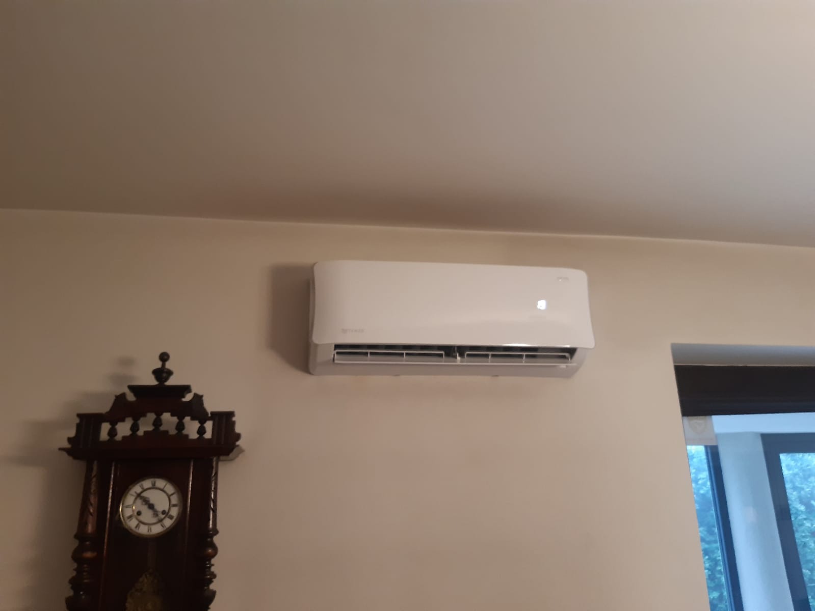 Biały klimatyzator ścienny zamontowany nad ozdobnym zegarem stojącym, widoczne okno w tle.