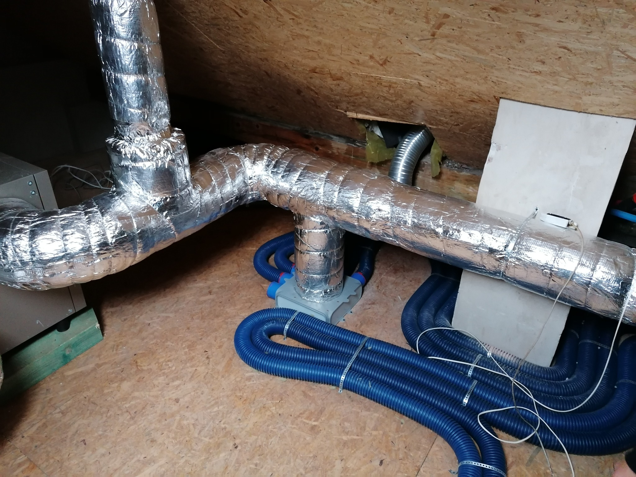 Rozbudowana instalacja wentylacyjna z niebieskimi rurami karbowanymi i izolowanymi przewodami aluminiowymi, widoczna w przestrzeni poddasza z drewnianą konstrukcją stropu.