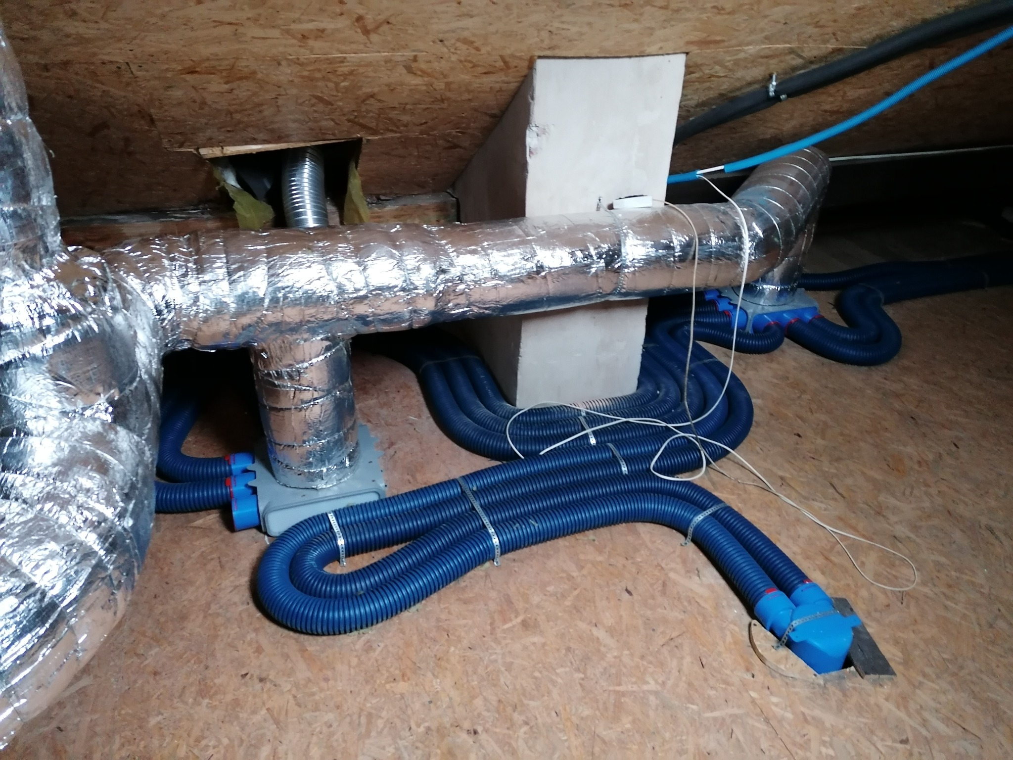 Instalacja wentylacyjna z rurami w kolorze niebieskim, zabezpieczona folią aluminiową, zamontowana na drewnianej podłodze strychu.