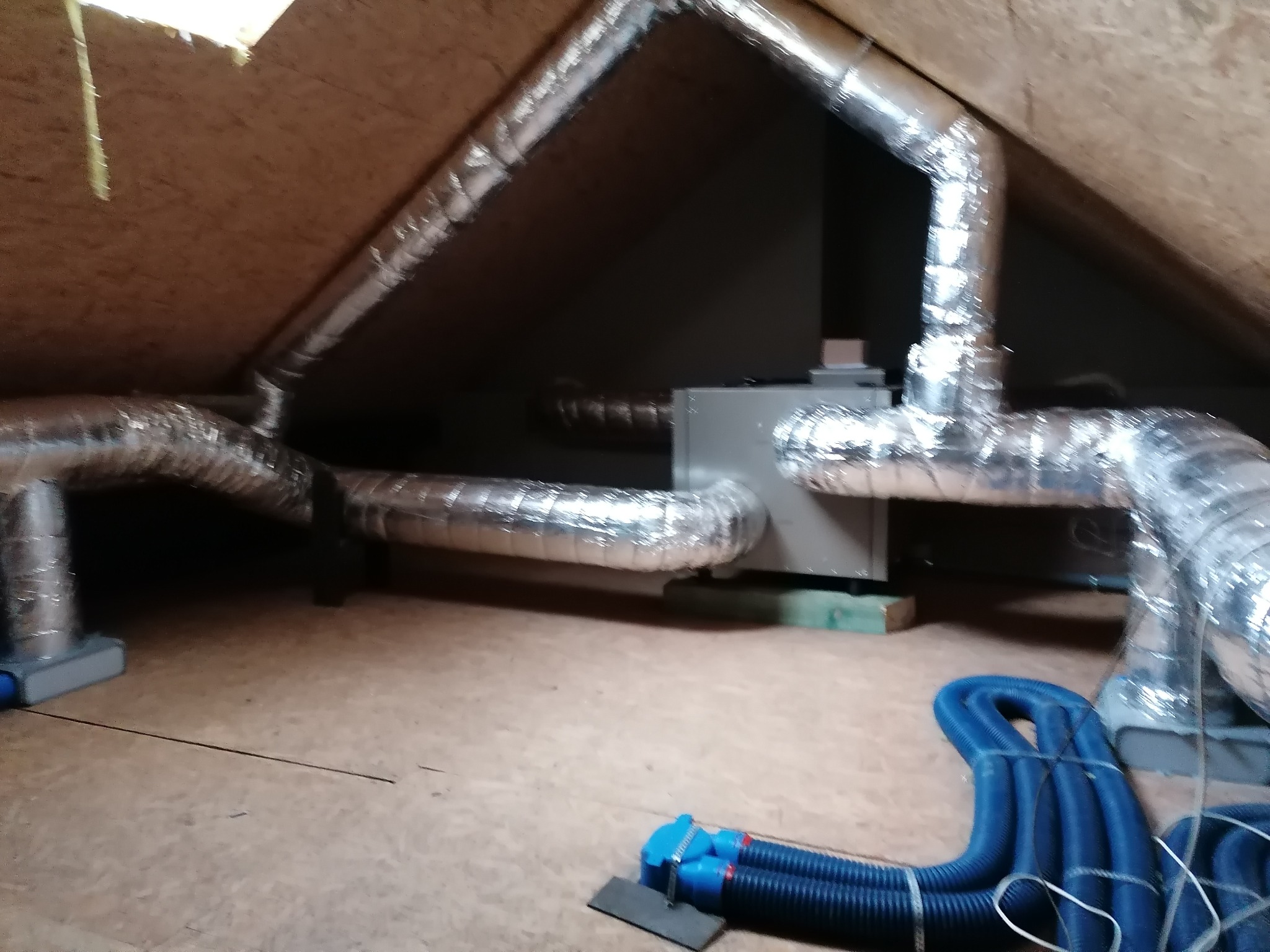 Instalacja wentylacyjna z aluminiowymi przewodami w przestrzeni poddasza, widoczny centralny rekuperator i niebieskie rury wentylacyjne na podłodze.