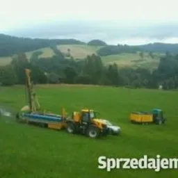 Żółty traktor z wiertnicą na przyczepie, w tle zielone wzgórza i las, widoczny proces wiercenia z rozpryskującą się wodą.