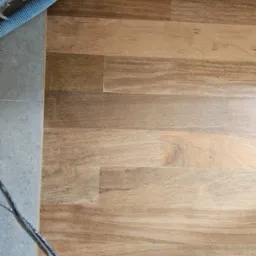 Fragment podłogi z drewnianych desek w trakcie renowacji, widoczny wąż od odkurzacza przemysłowego i przewody, szlifowana powierzchnia z widocznymi słojami.