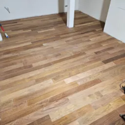 Ułożona drewnopodobna podłoga w jasnym pomieszczeniu z białymi ścianami, widoczny wałek malarski przy ścianie i zwinięty kabel elektryczny od maszyny do obróbki podłóg w prawym dolnym rogu...