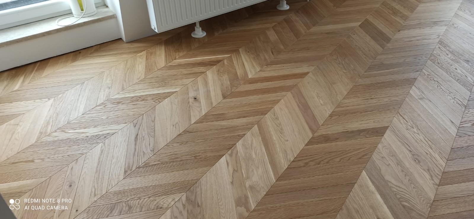 Ułożony parkiet w jodełkę w Żerkowie, widoczny fragment okna i grzejnika, jasne drewno.