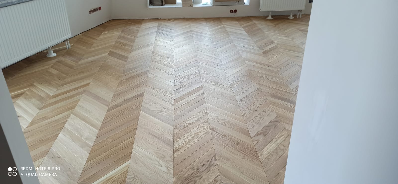 Jasny parkiet ułożony w jodełkę francuską, widoczne grzejniki i fragment ściany w trakcie malowania.