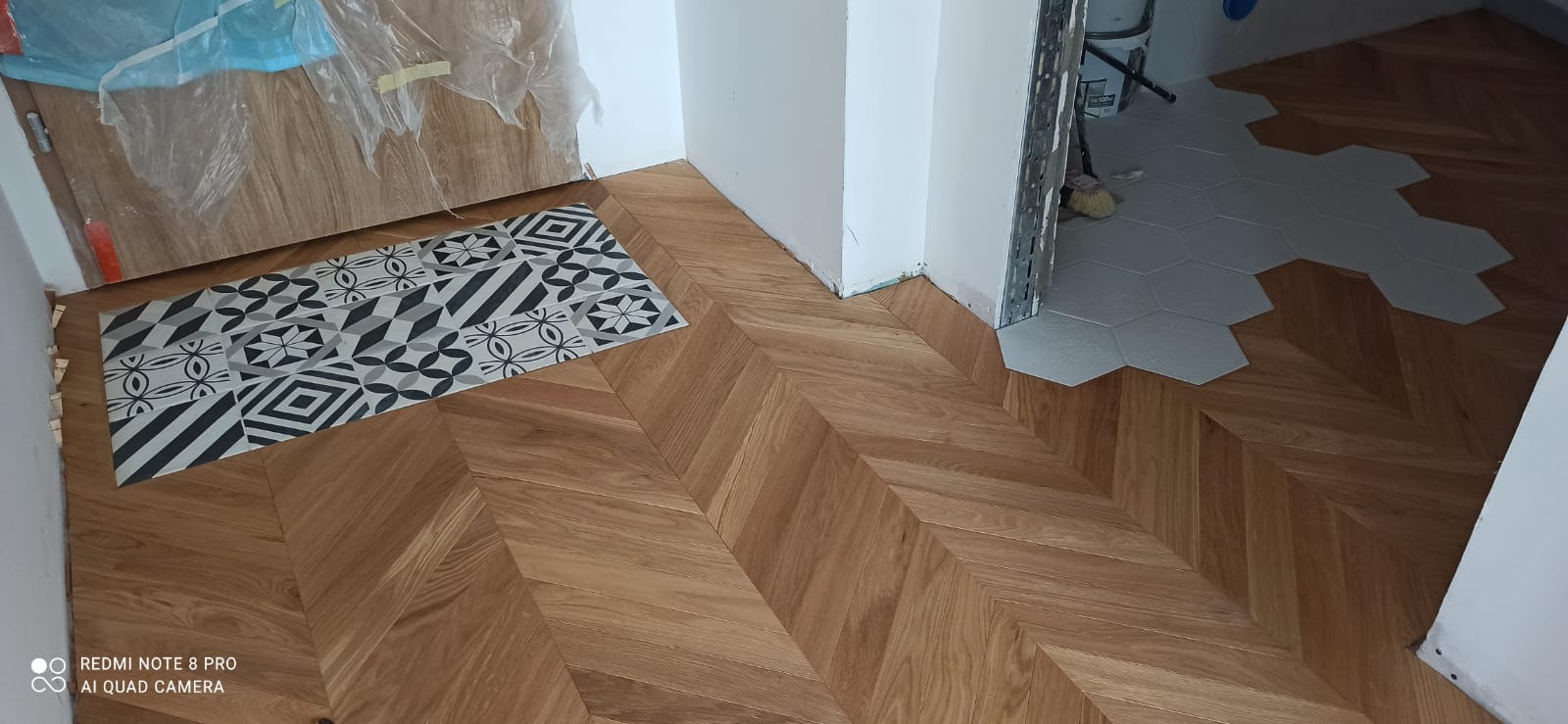 Ułożony parkiet w jodełkę przechodzi w heksagonalne płytki podłogowe w progu, z fragmentem dekoracyjnej płytki przy drzwiach, widoczne ślady po remoncie.