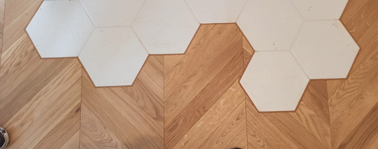 Jasny parkiet ułożony we wzór jodełki francuskiej, przeplatany białymi, sześciokątnymi płytkami ceramicznymi.