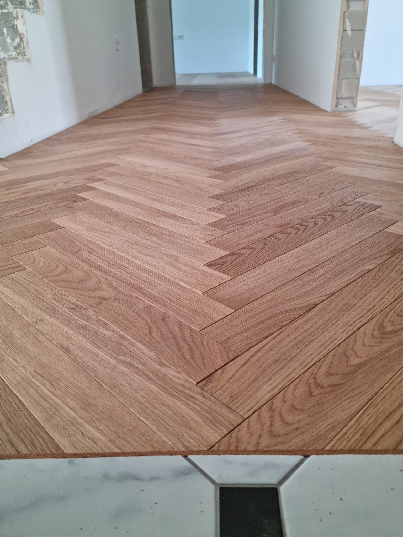 Ułożony parkiet w jodełkę, z widocznymi niedokończonymi fragmentami ścian i przejściem do innego pomieszczenia w tle.