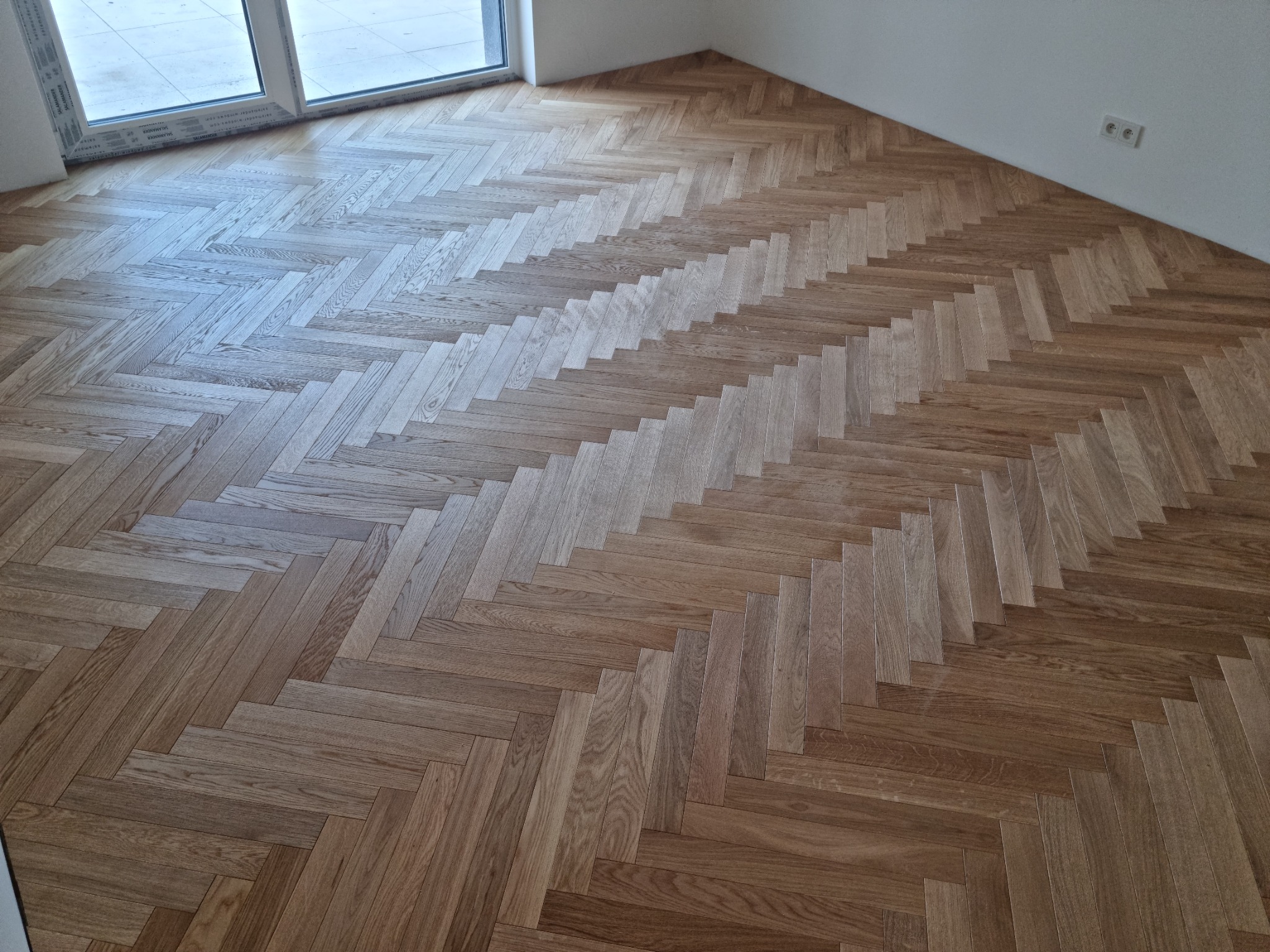 Ułożony parkiet w jodełkę francuską, z widoczną strukturą drewna i wykończonymi listwami przypodłogowymi, oświetlony naturalnym światłem wpadającym przez okno balkonowe.