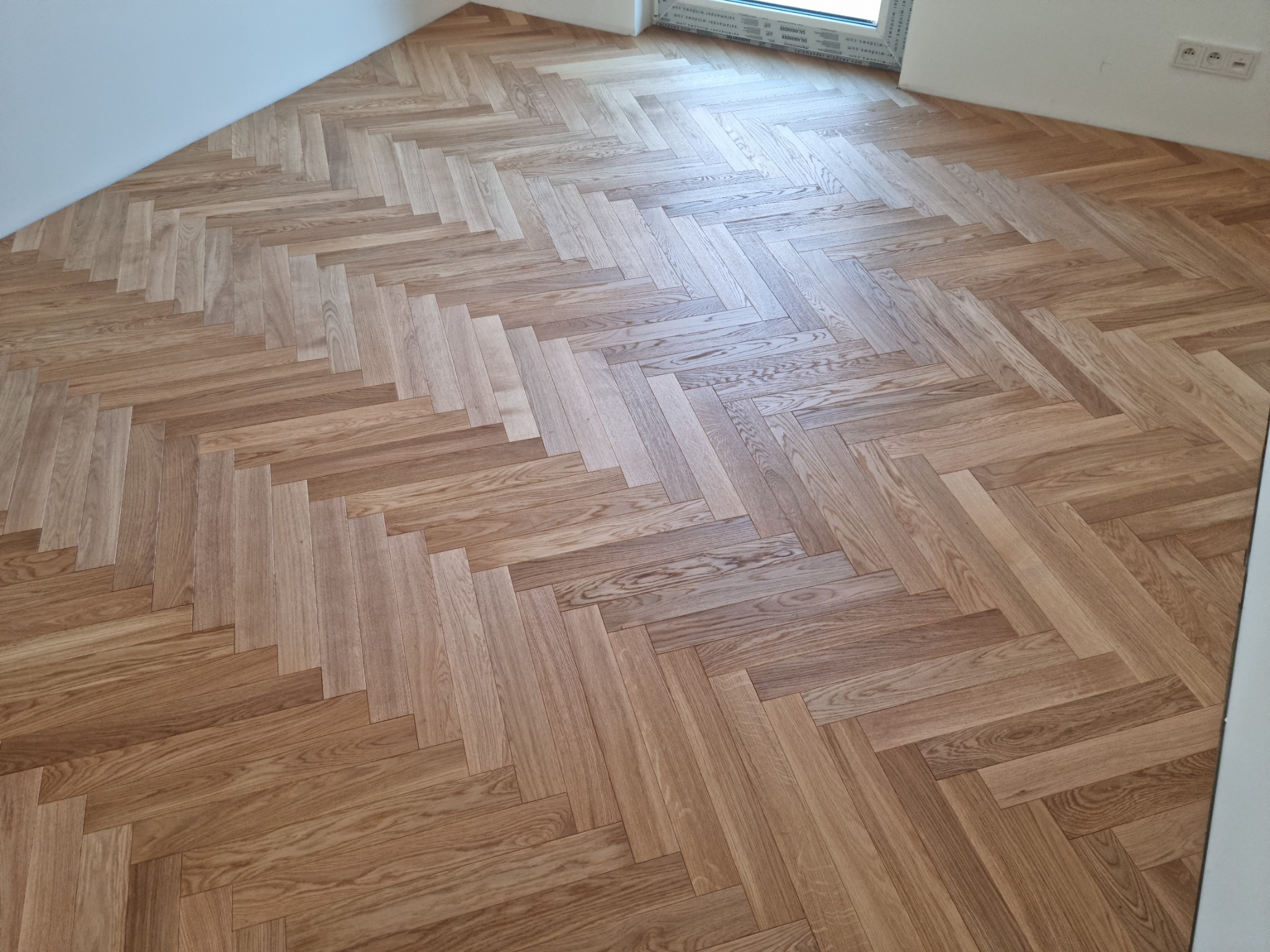 Ułożony parkiet dębowy w jodełkę francuską, widoczne naturalne usłojenie drewna, białe ściany i okno w tle.