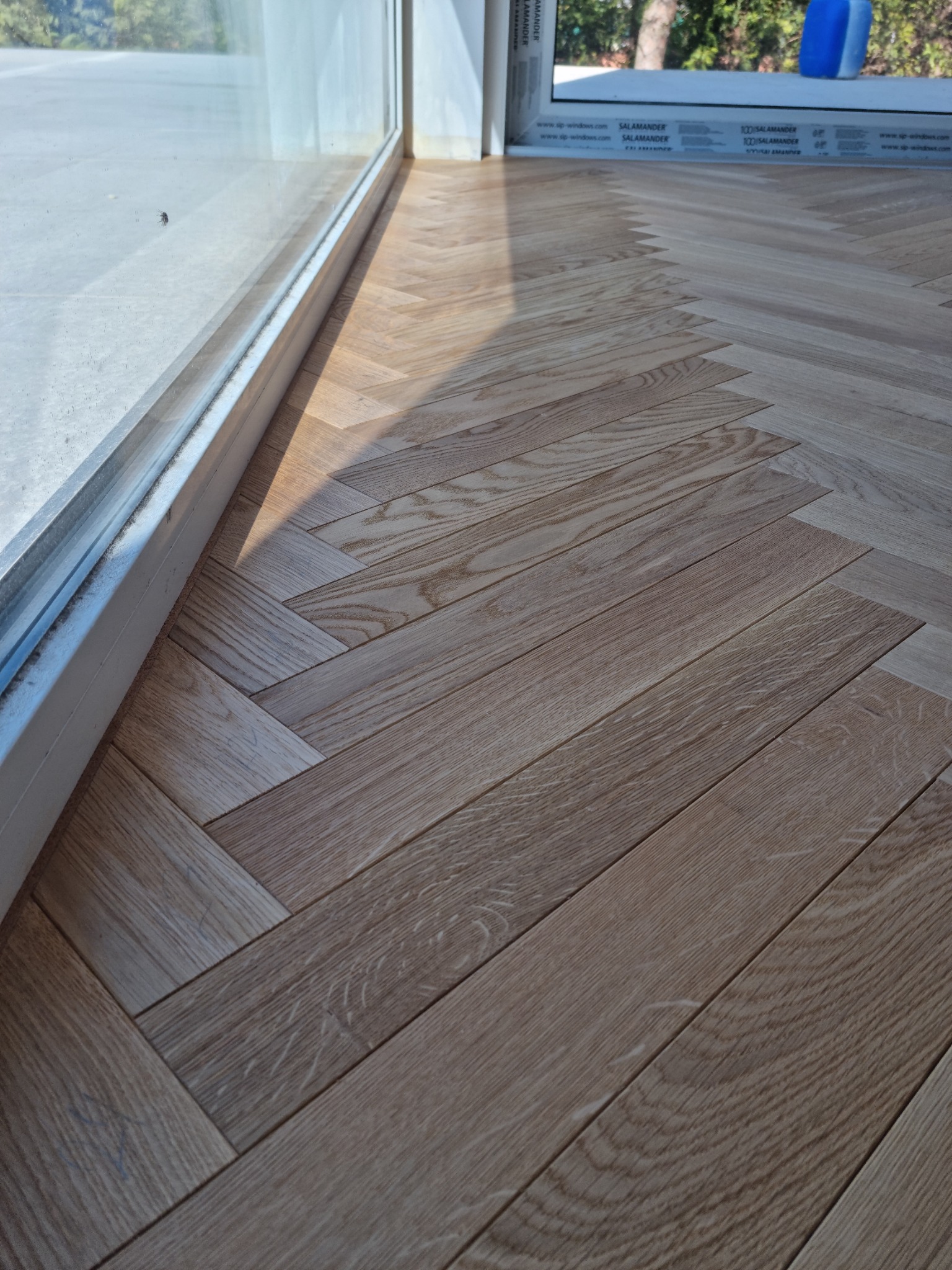 Ułożony parkiet w jodełkę przy oknie, widoczne detale słojów drewna i cień padający na podłogę.