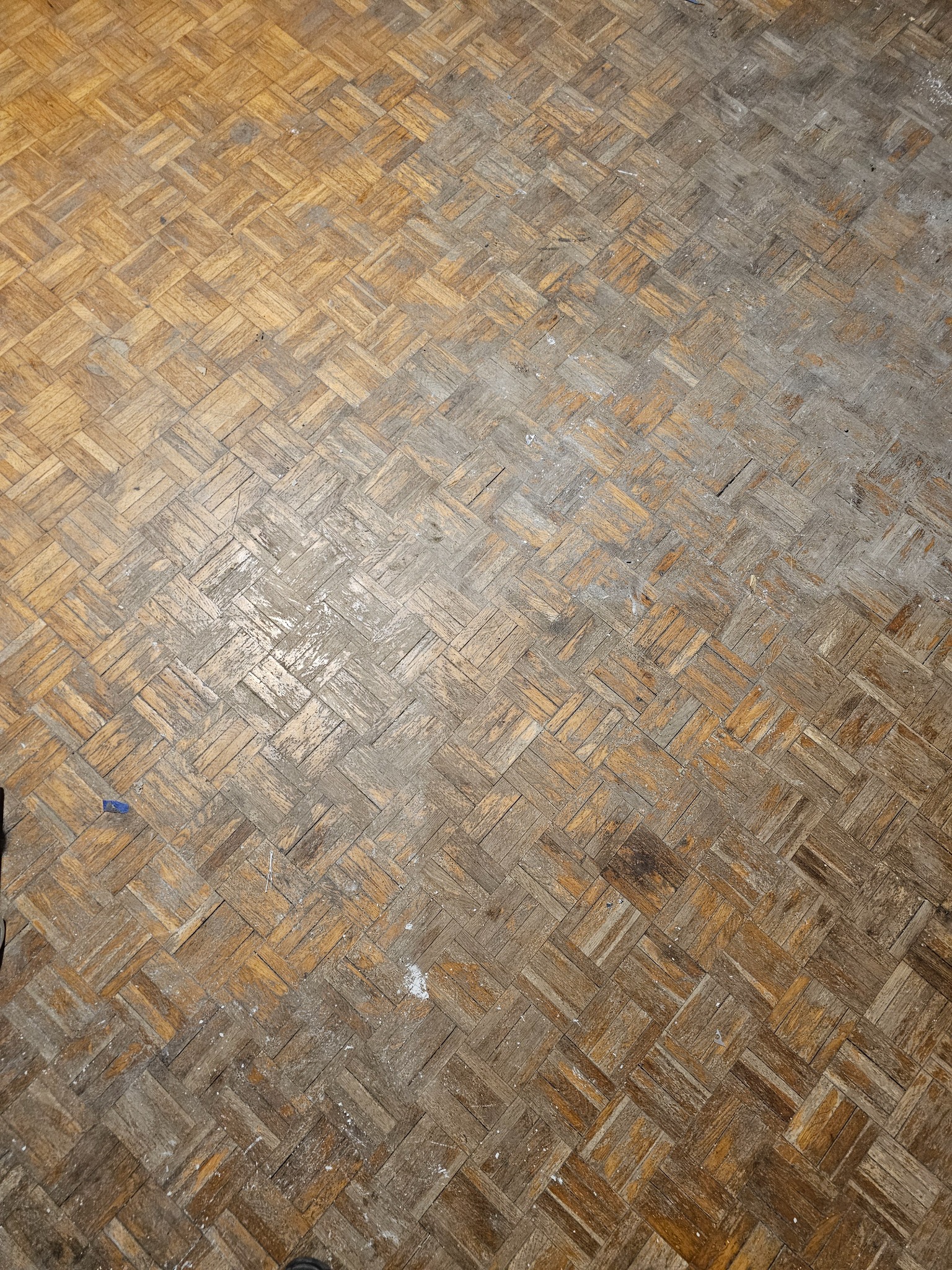 Zużyta, mozaikowa podłoga parkietowa z widocznymi śladami użytkowania, rysami i zabrudzeniami, ułożona w charakterystyczny wzór koszykowy.