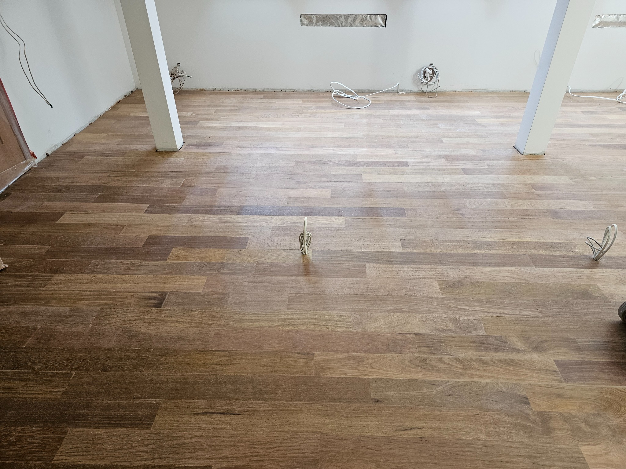 Nowo ułożona podłoga z parkietu w pustym pomieszczeniu z białymi ścianami i słupami nośnymi, widoczne kable elektryczne wystające ze ścian.