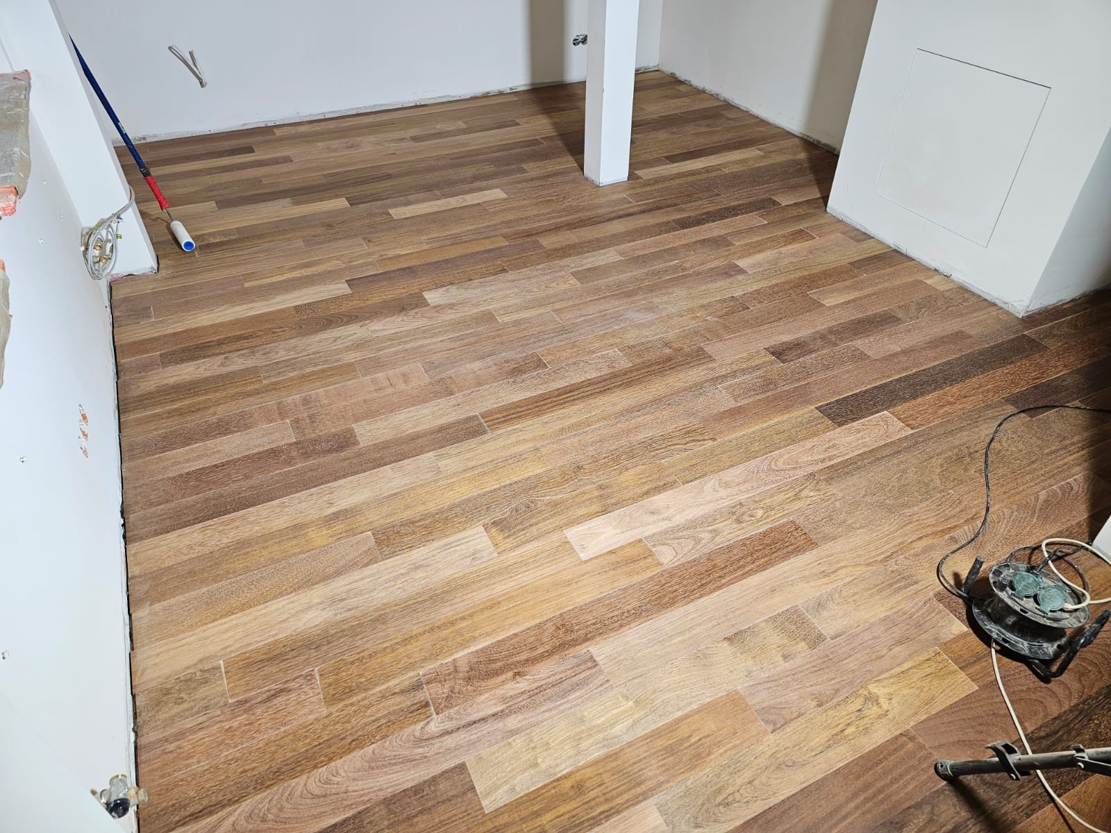 Świeżo ułożony parkiet w odcieniach brązu i beżu, z widoczną maszyną do polerowania w rogu pomieszczenia i narzędziami malarskimi przy ścianie.