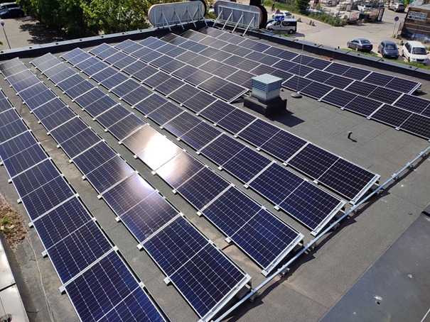 Rząd paneli słonecznych na płaskim dachu budynku, z widoczną wentylacją i fragmentem otoczenia miejskiego w tle.