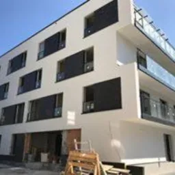 Nowoczesny, trzypiętrowy budynek z białymi ścianami i ciemnymi okładzinami okiennymi. Widoczne balkony z szklanymi balustradami. Część elewacji na parterze w trakcie wykończenia, widoczne...