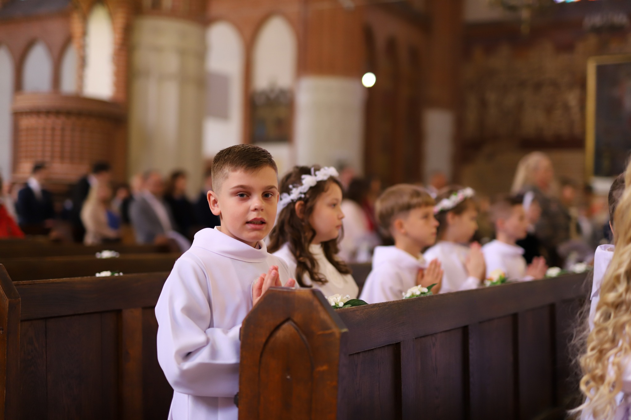 Chłopiec w białej albie w kościele, z rękami złożonymi do modlitwy. W tle inne dzieci w strojach komunijnych i wierni w ławkach. Uroczysty nastrój.