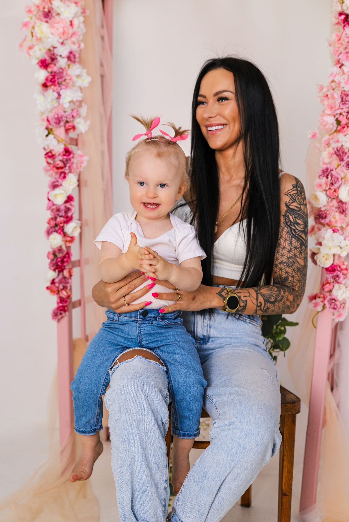 Uśmiechnięta mama z tatuażami trzyma na kolanach roześmianą blondwłosą córeczkę na tle dekoracji z różowych kwiatów.