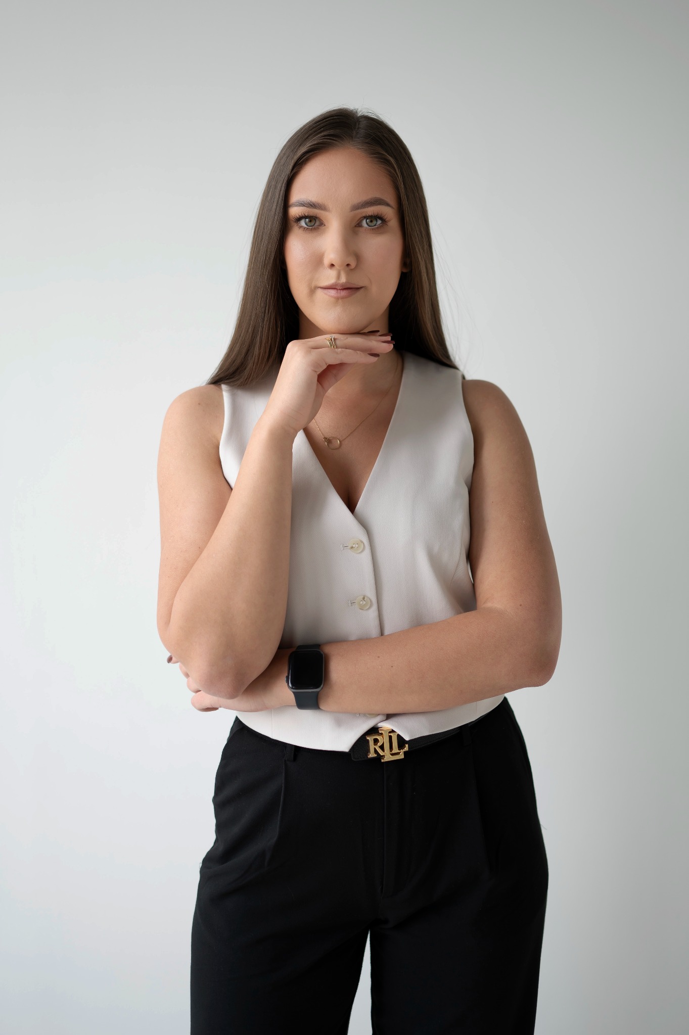 Portret kobiety w białej kamizelce i czarnych spodniach, z ręką pod brodą i zegarkiem na nadgarstku, na jasnym tle. Złoty pierścionek i naszyjnik.