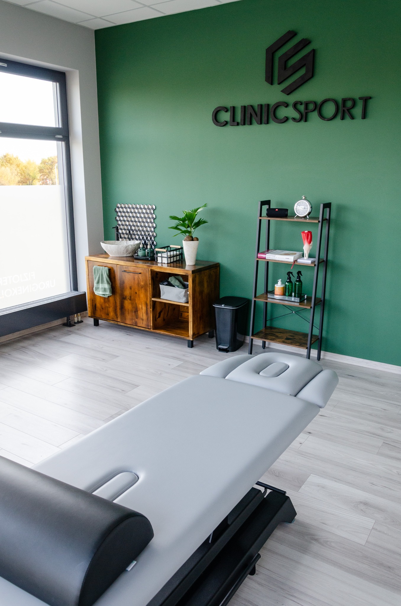 Jasne wnętrze gabinetu z szarą leżanką, drewnianą komodą z umywalką i półkami, zielona ściana z logo Clinic Sport.