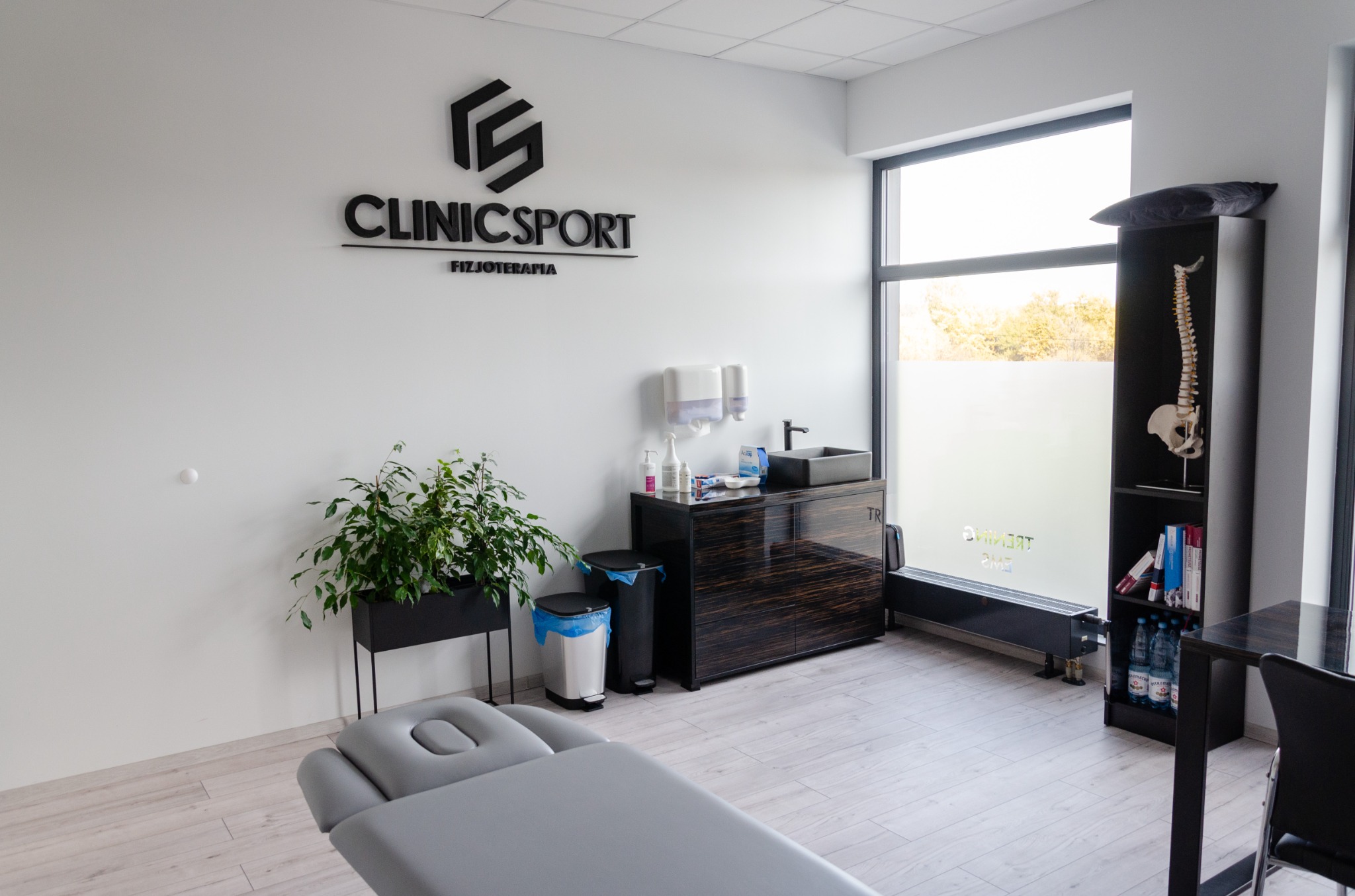 Jasne wnętrze gabinetu z leżanką, rośliną w czarnej donicy, szafką z umywalką i logo 'ClinicSport Fizjoterapia' na ścianie, model kręgosłupa na półce.