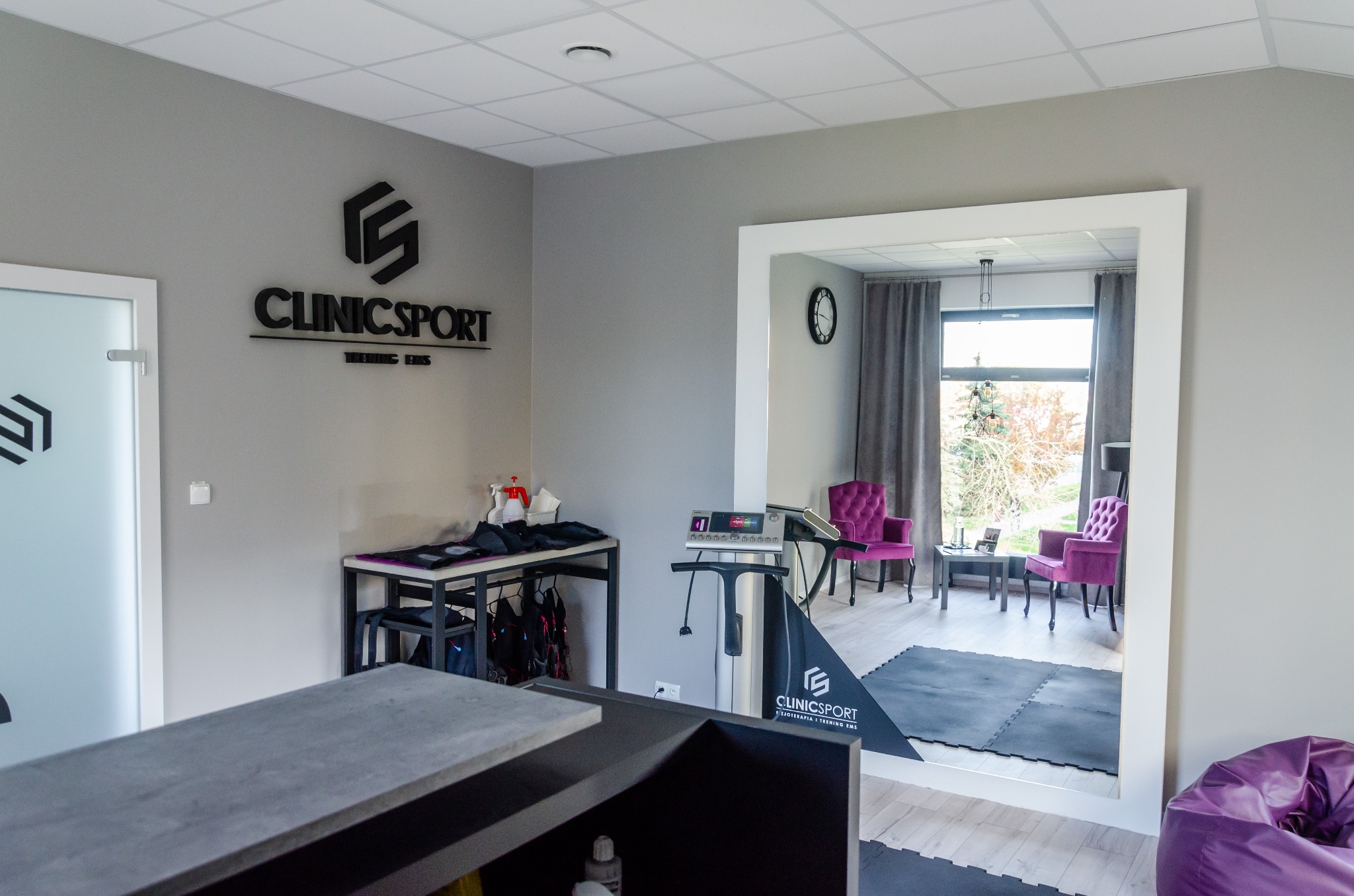 Wnętrze gabinetu z recepcją, lustrem odbijającym salę ćwiczeń z fioletowymi fotelami, urządzeniem do ćwiczeń, logo firmy ClinicSport na ścianie i drzwiach.