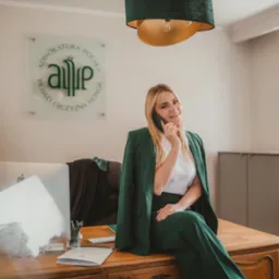 Kobieta w zielonym garniturze siedzi na biurku w gabinecie, rozmawiając przez telefon; w tle widoczne logo kancelarii i nowoczesny monitor.