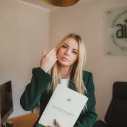 Kobieta w zielonym garniturze, siedząca przy biurku, trzymająca folder z logo kancelarii adwokackiej i danymi adwokata, w tle lampa i logo kancelarii.